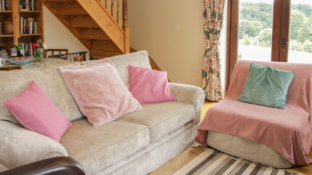 A living room with a beige sofa, a wooden staircase, a bookshelf, and a view through a window. - Minton Lane Cottage (Church Stretton)