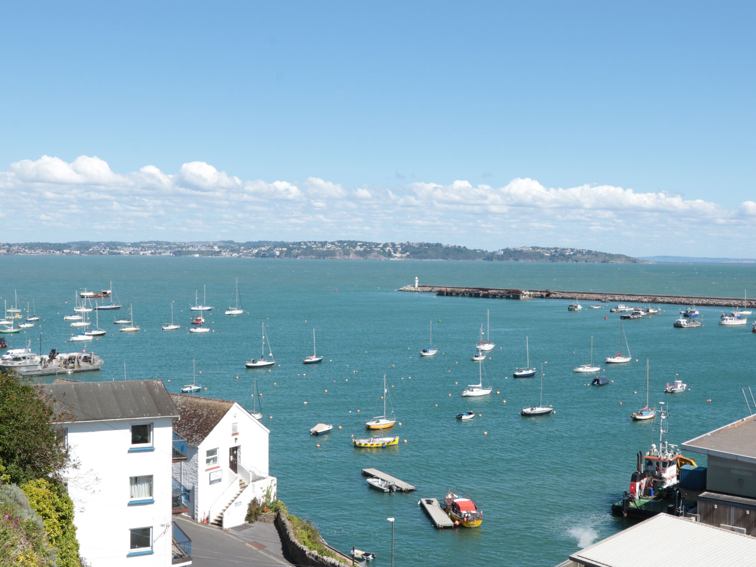 A harbor with multiple boats docked, a lighthouse in the distance, and a white building with a balcony overlooking the water.