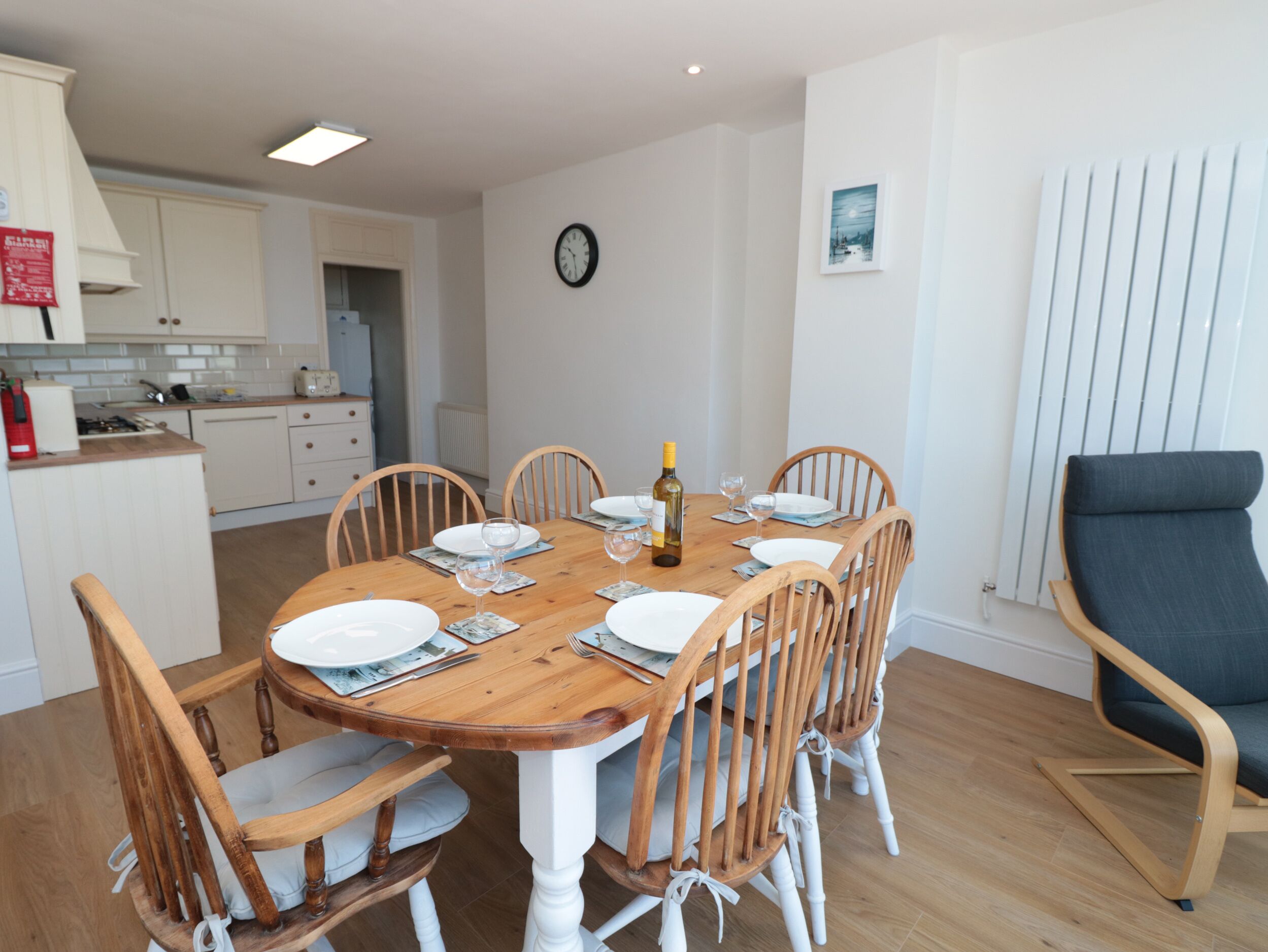 Un coin repas avec une table en bois dressée pour six personnes, entourée de chaises en bois dotées d’assises rembourrées. Une horloge et un tableau encadré sont accrochés au mur.