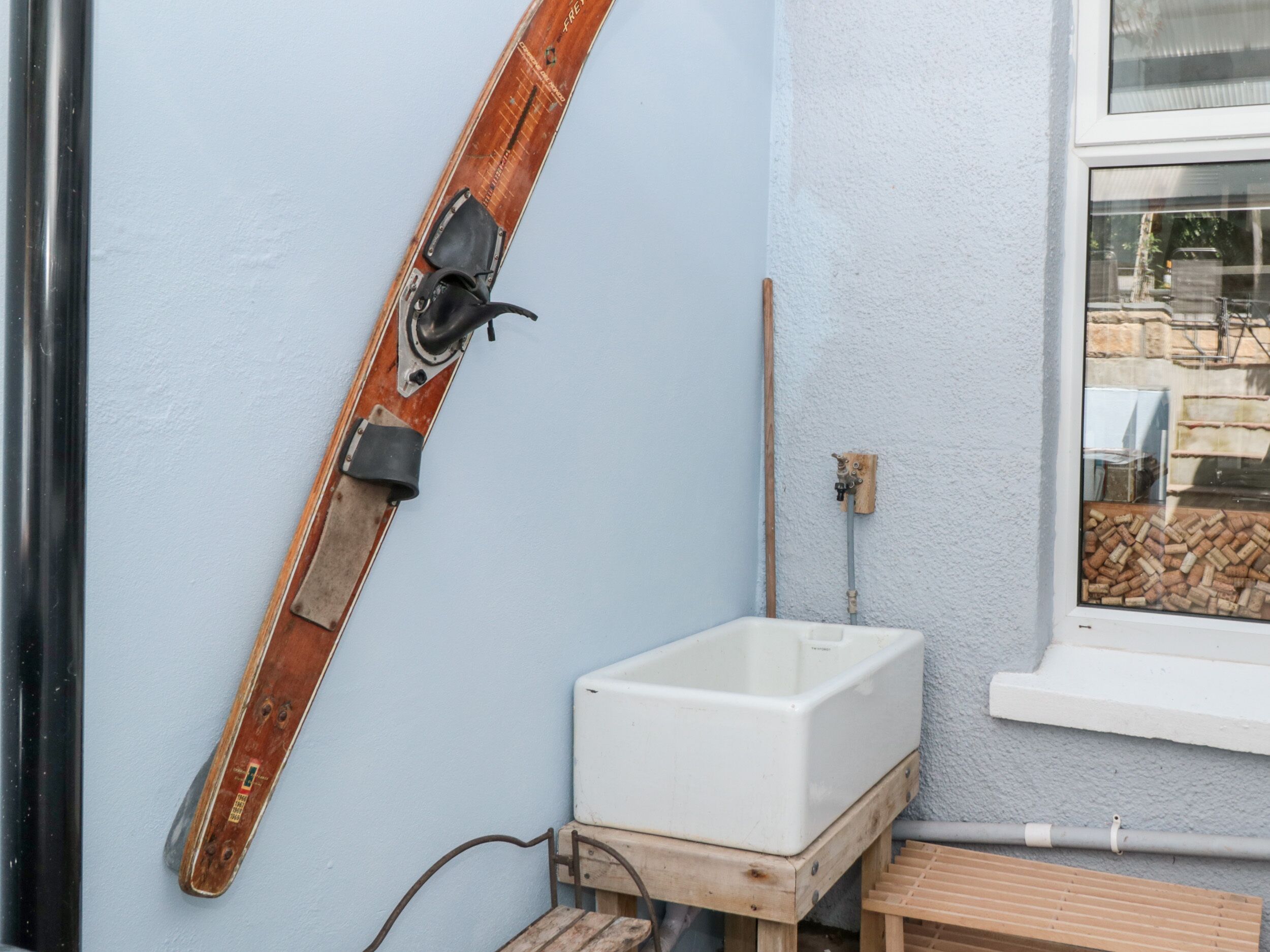 A bathroom with a wooden ski leaning against the wall, a white basin, and a window with a view of outdoor furniture.
