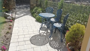 Un patio empedrado con una mesa redonda y sillas, rodeado de vegetación y un muro de piedra.