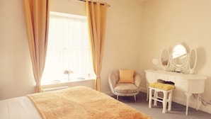 Un dormitorio con cama, una silla, un tocador con espejos y una ventana con cortinas.