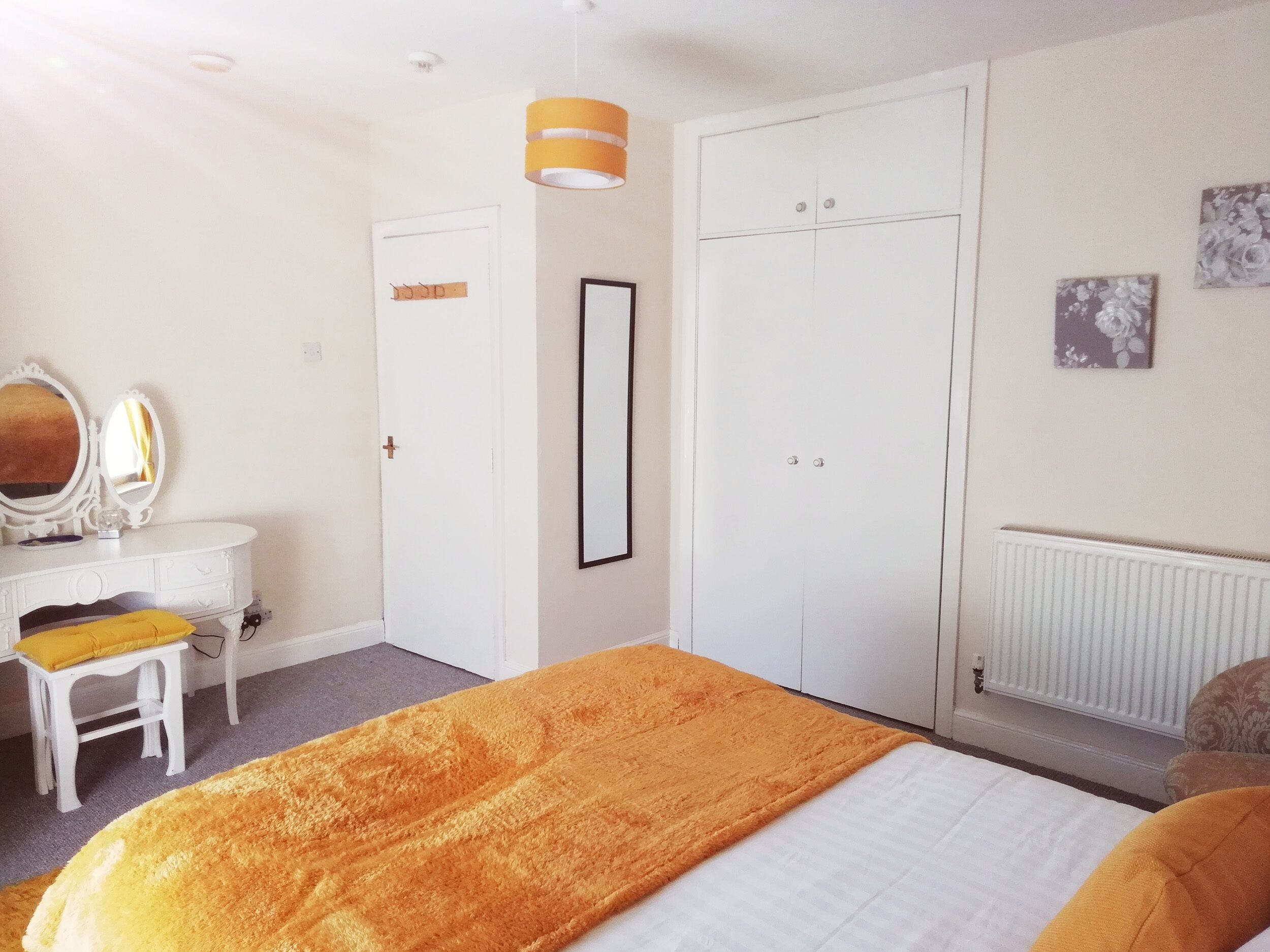 Un dormitorio con una cama, un aparador blanco con espejo, una puerta y un radiador.