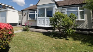 A two-story house with a white exterior, a brown roof, and a small garden with pink flowers and a green bush. - Garden View (Newquay)