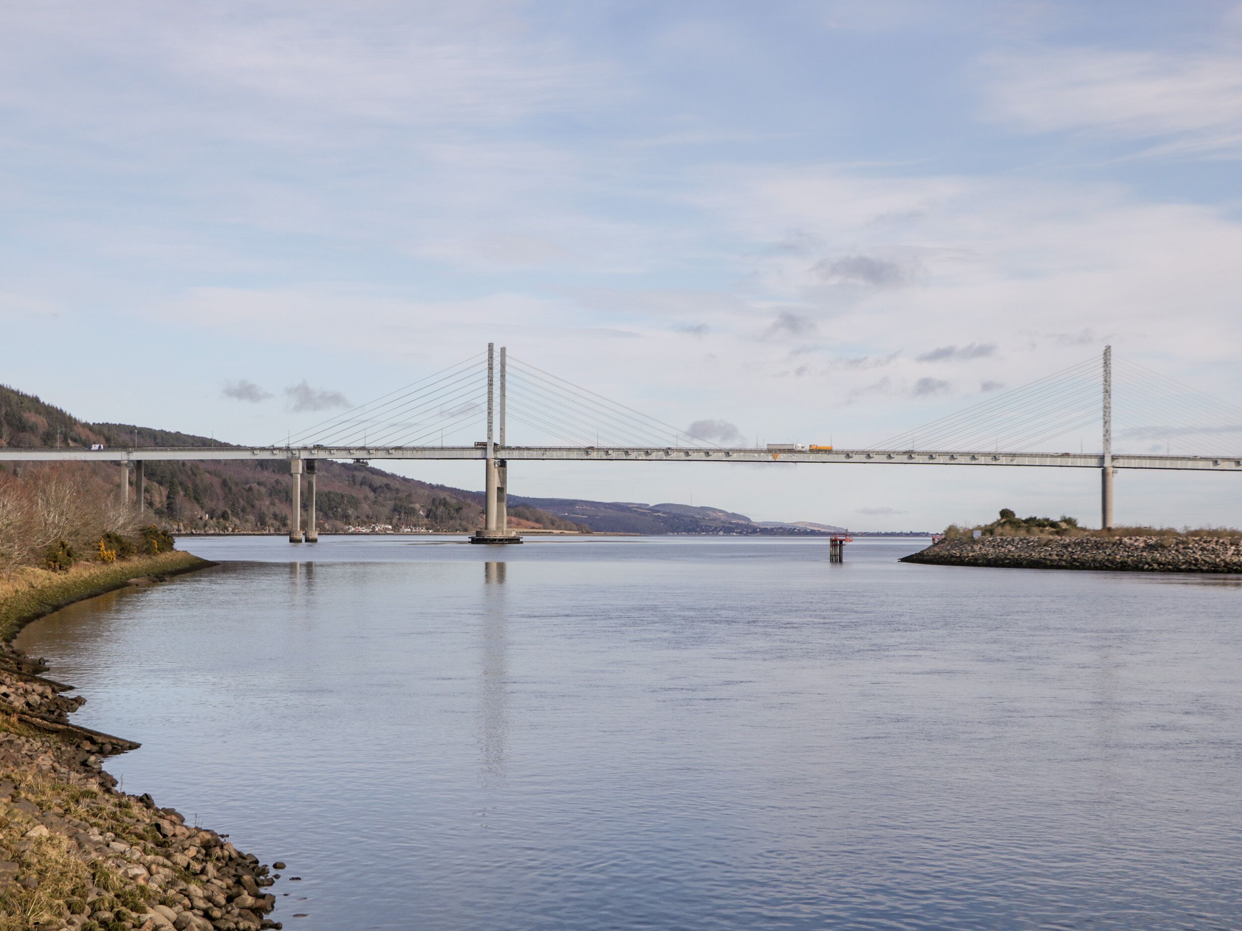 A suspension bridge over a calm river with rocky banks and hills in the background.