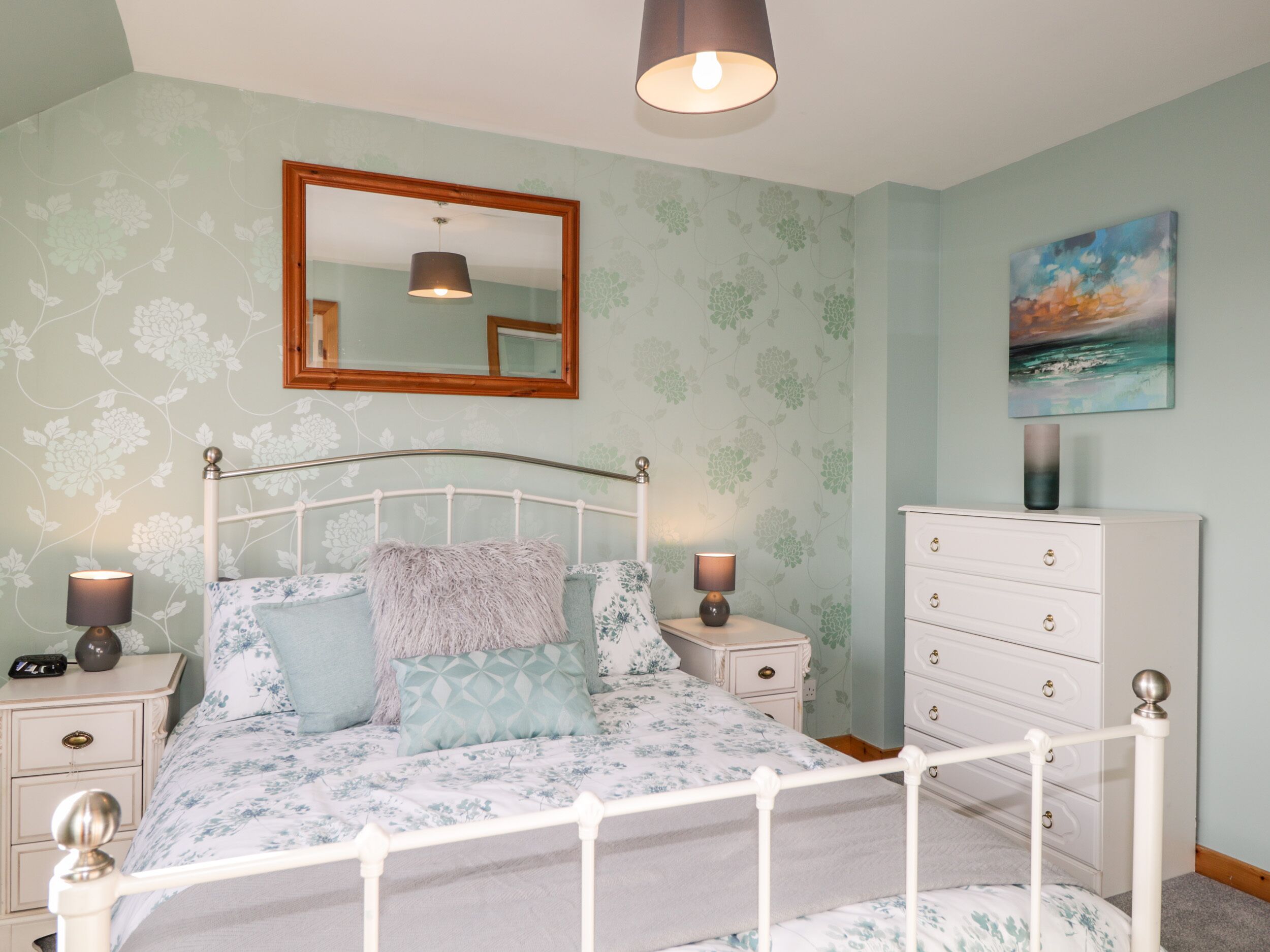 A bedroom with a white metal bed frame, floral-patterned bedding, two bedside lamps, a white dresser, and a painting of a seascape.