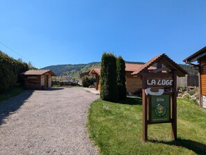 Ländliche Szenerie mit einem Schotterweg, der zu Holzhäusern führt, einem Schild für 'La Luge Gîte' und einem Berg im Hintergrund.