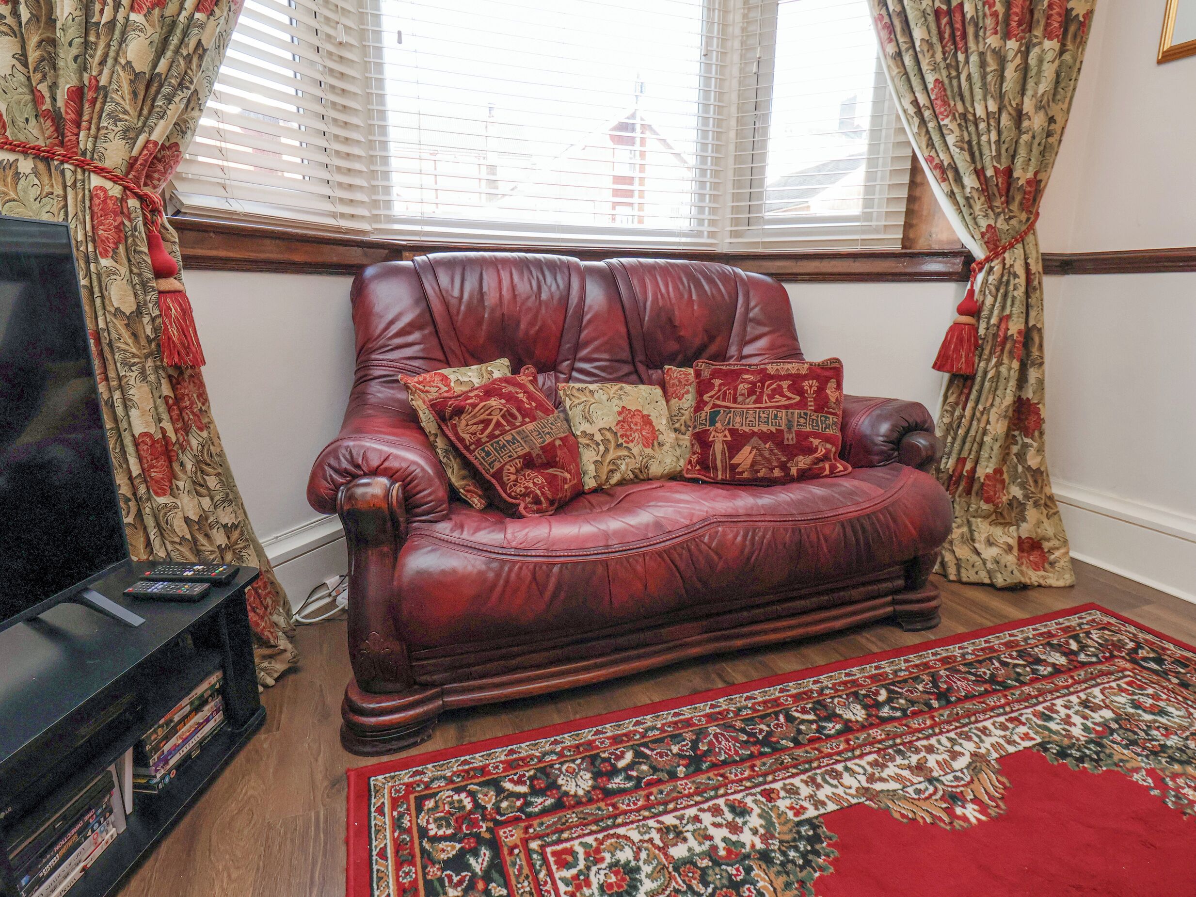 A leather sofa with floral cushions, a television, and a patterned rug in a room with wooden flooring and floral curtains.