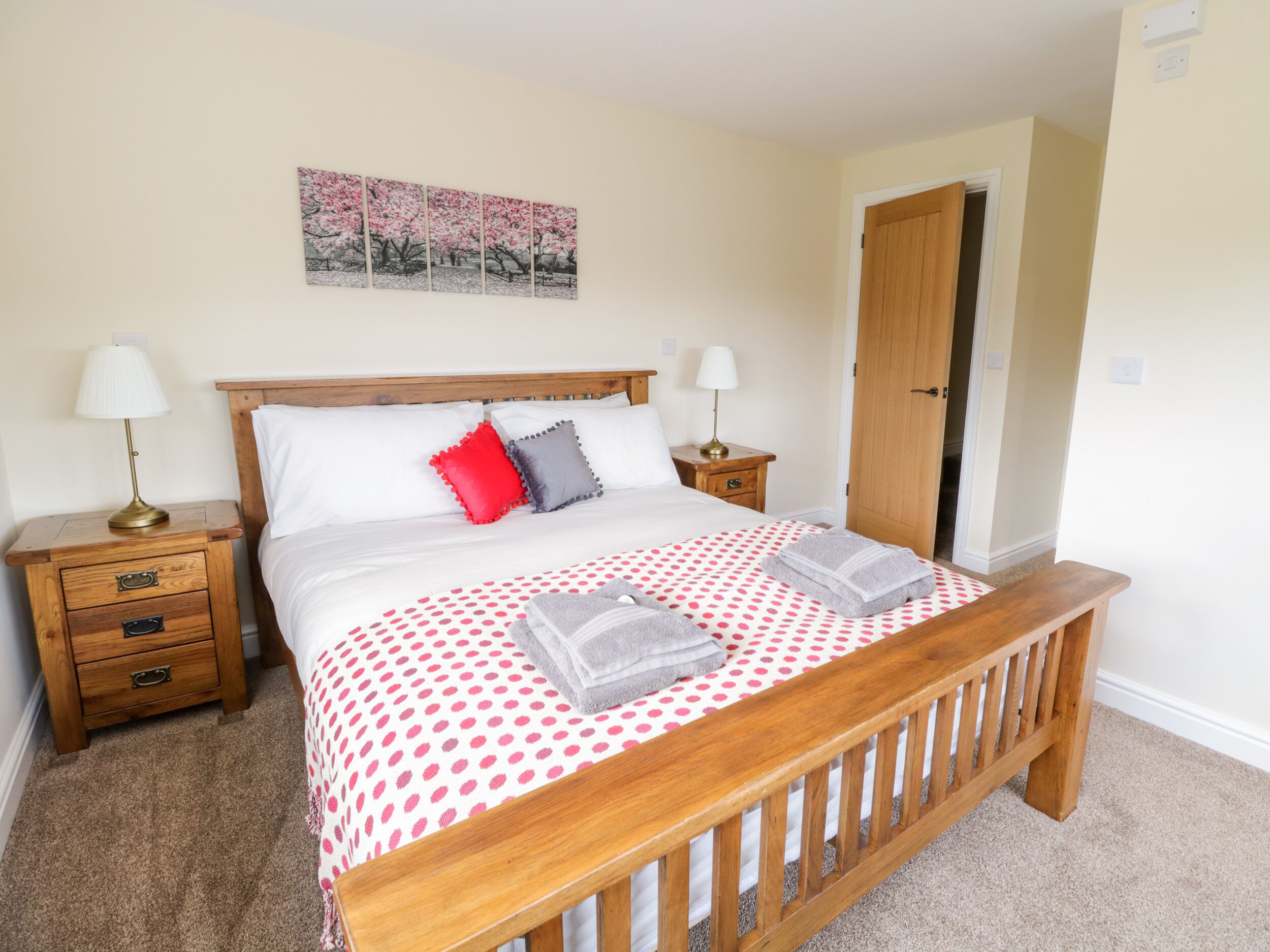A bedroom with a wooden bed, two bedside tables, a nightstand, a red and grey pillow, a polka dot bedspread, and a painting on the wall.