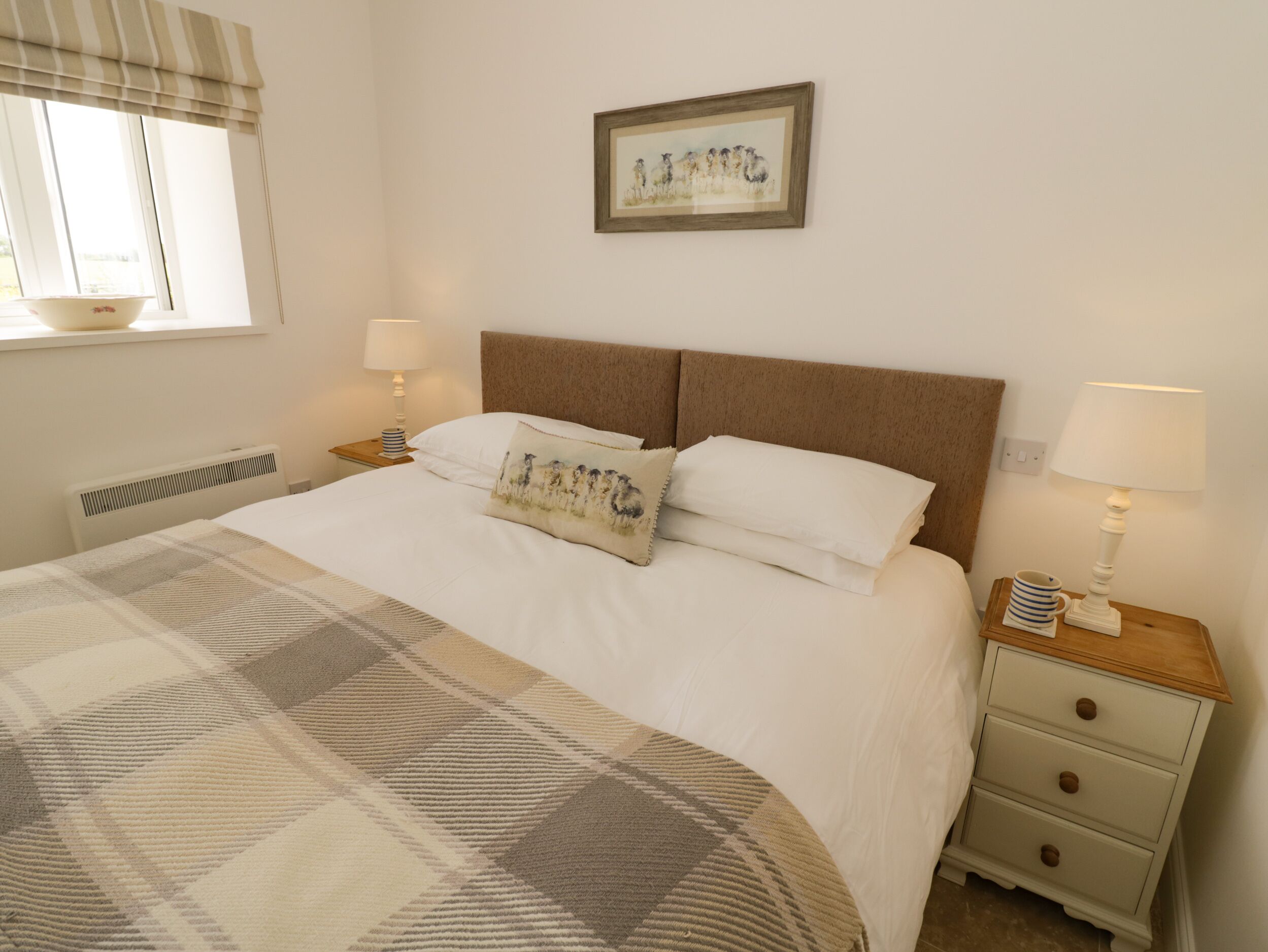 A neatly made bed with a plaid blanket, two bedside lamps, a framed picture on the wall, and a window with a curtain.