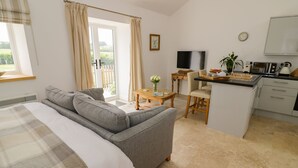 A modern hotel room with a large bed, a grey sofa, a small dining table, a kitchen area, and a sliding glass door leading to a balcony.