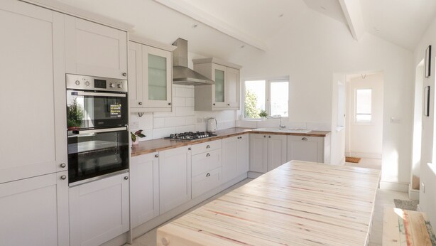 A modern kitchen with white cabinetry, a built-in oven, a gas stove, and a wooden dining table. - Y Gogarth (Tyn-y-Gongl)