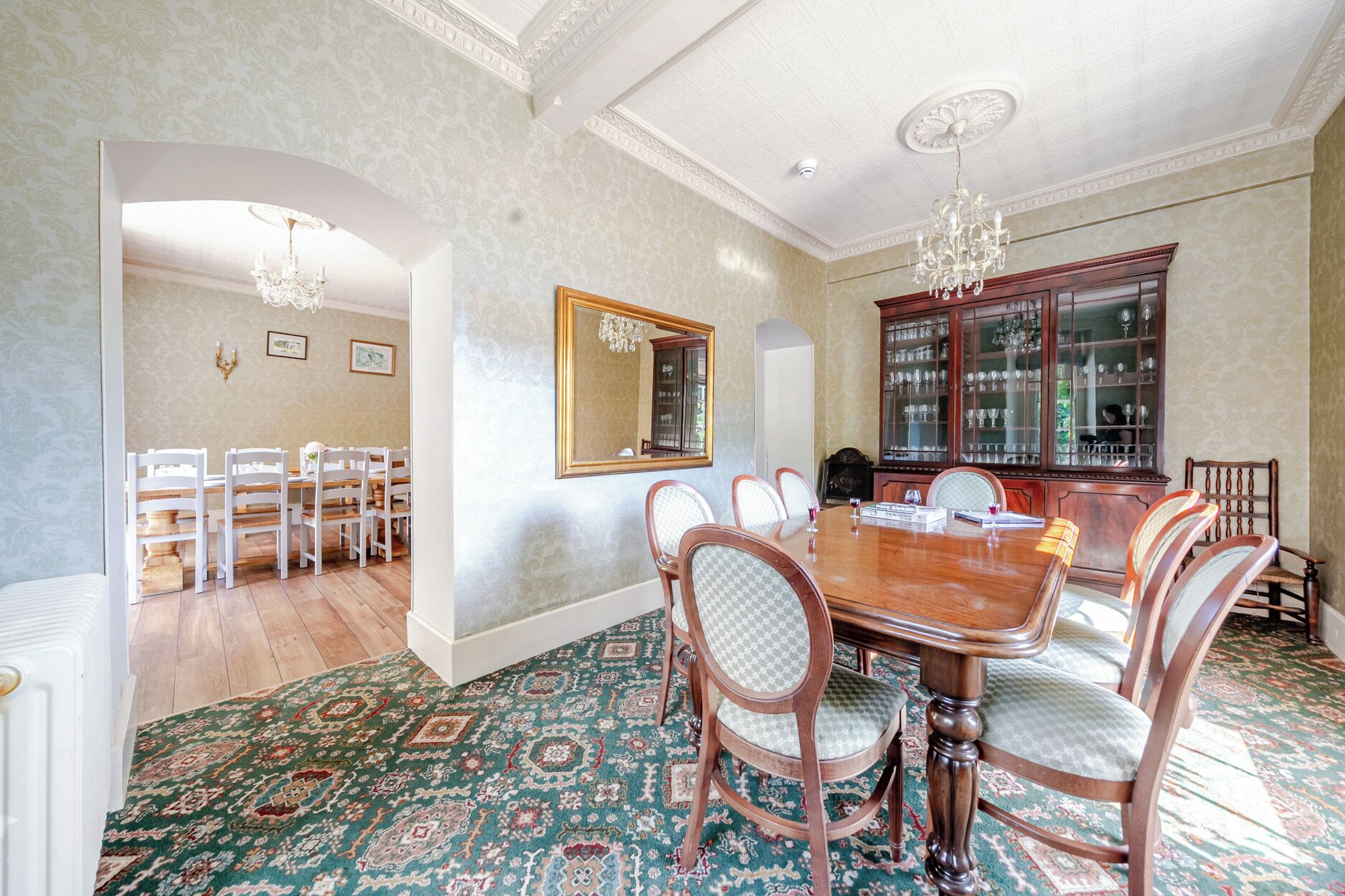A dining room with a large wooden table, chairs, a chandelier, a mirror, and a glass cabinet.