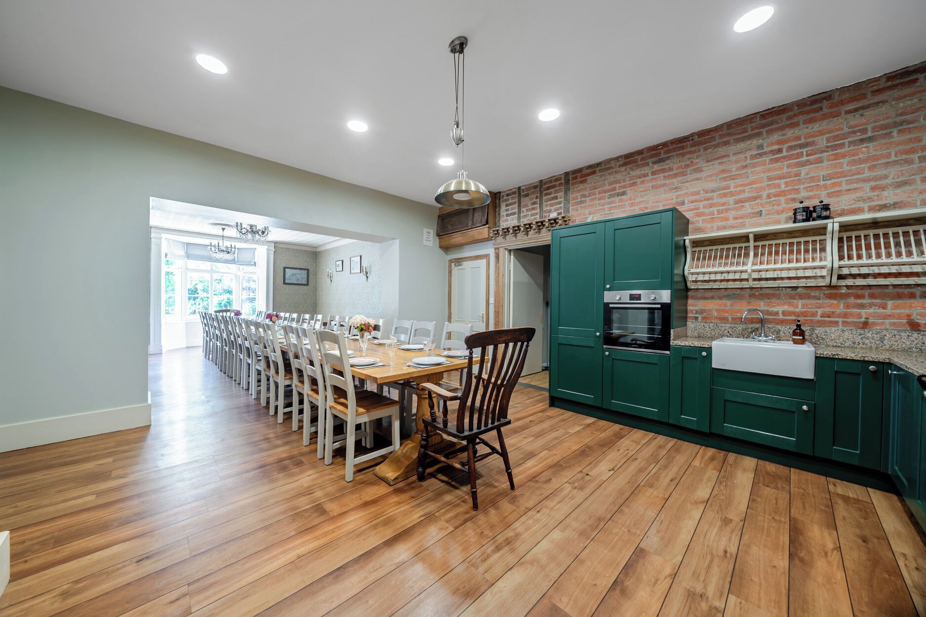 A dining area with a long wooden table and chairs, a kitchen with green cabinets and a brick wall, and a chandelier.