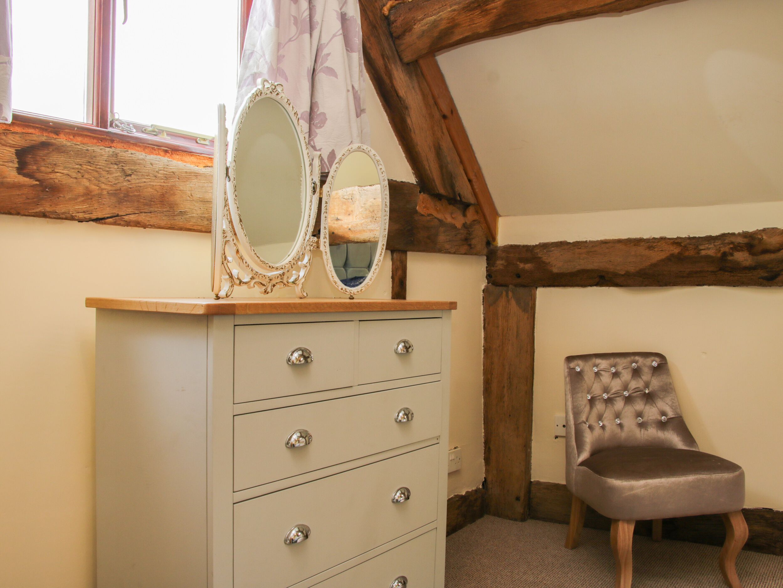 A room with a wooden ceiling, a beige dresser with three drawers, two mirrors, and a tufted chair.