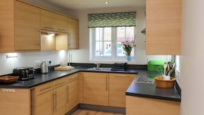 A kitchen with wooden cabinets, a countertop, a window with a view, and a vase with flowers. - Driftwood (Filey)