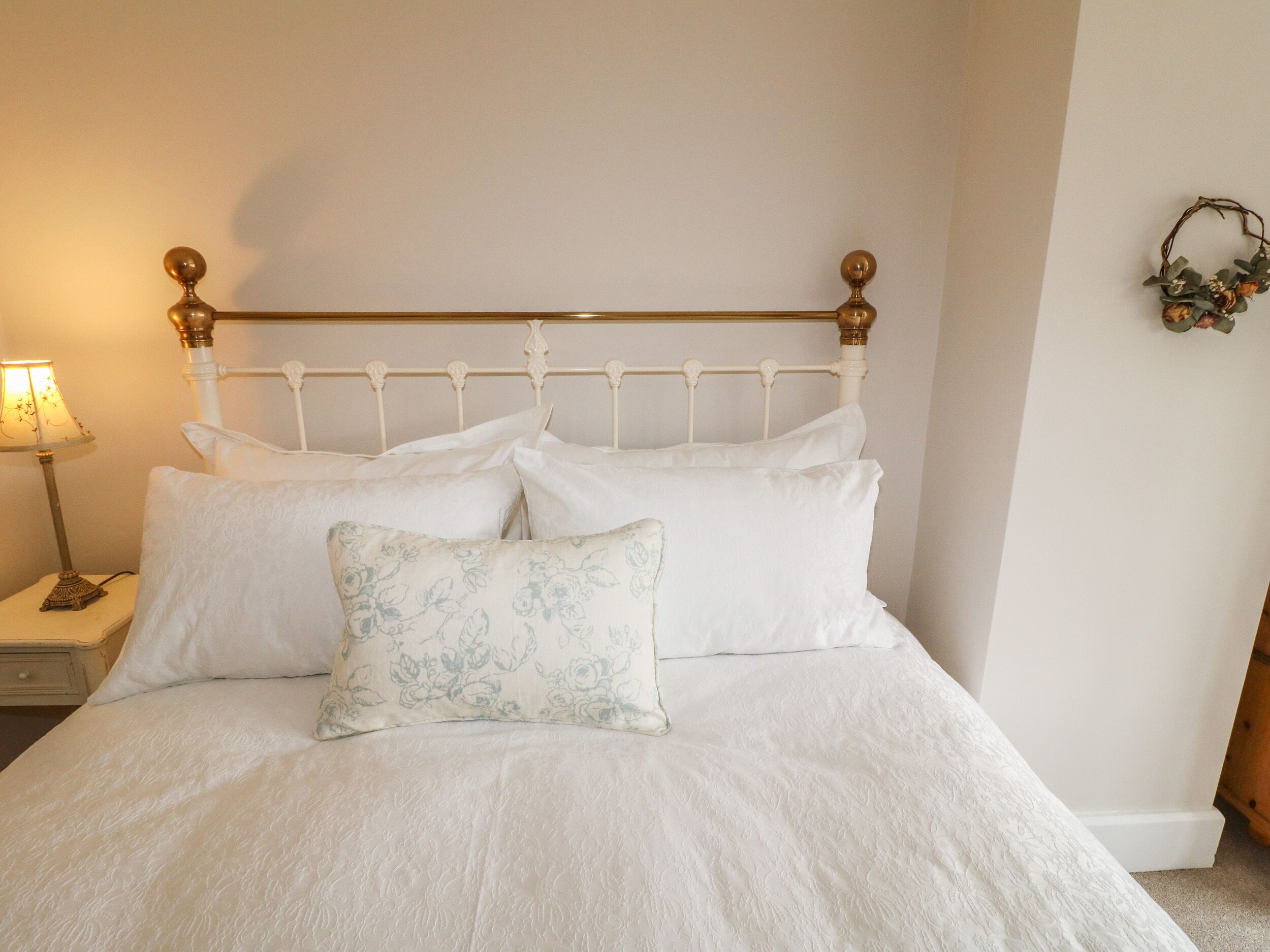 A neatly made bed with white linens and a floral pillow, a white metal headboard, and a bedside table with a lamp.