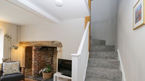 A living room with a fireplace, a television on a white stand, a chair with a cushion, and a staircase with a white railing. - Troopers Cottage (Frome)