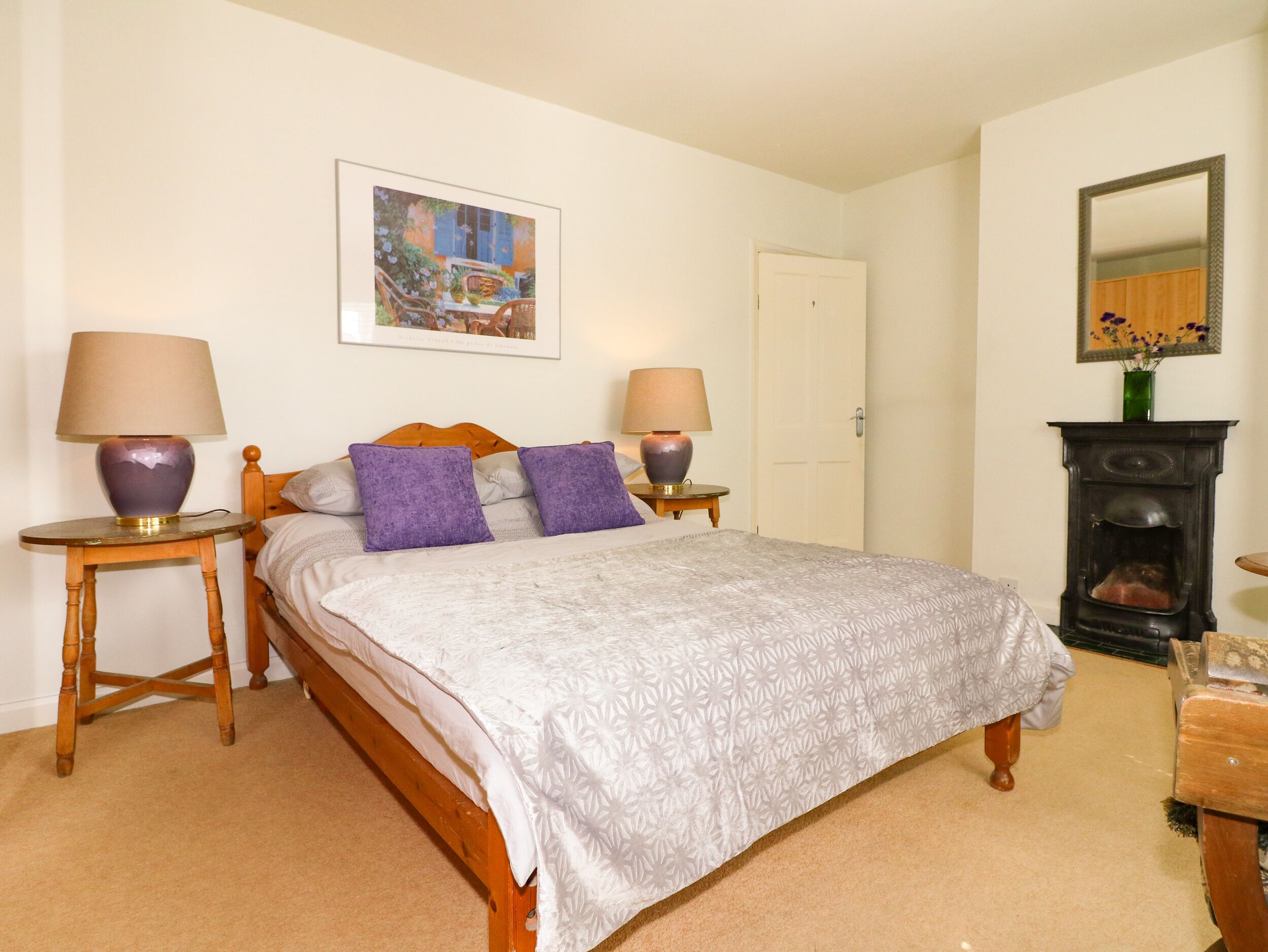 A bedroom with a bed, two bedside tables with lamps, a painting on the wall, a door, and a fireplace.