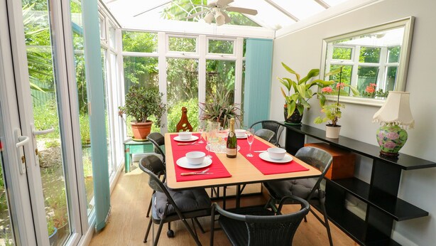 Comedor estilo invernadero con una mesa dispuesta para cuatro personas, sillas alrededor, ventilador de techo y plantas en macetas.