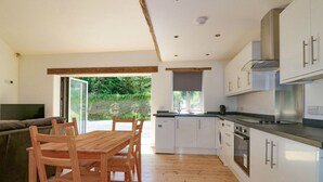A modern kitchen with wooden flooring, white cabinets, and a dining area with a wooden table and chairs. - The Cart House (St Austell)