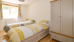 A bedroom with two beds, a wardrobe, a radiator, and a window with curtains. - The Cart House (St Austell)