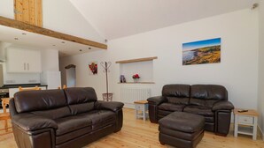 A spacious living room with a brown leather sofa and ottoman, a wooden dining table, and a kitchen in the background. - The Cart House (St Austell)