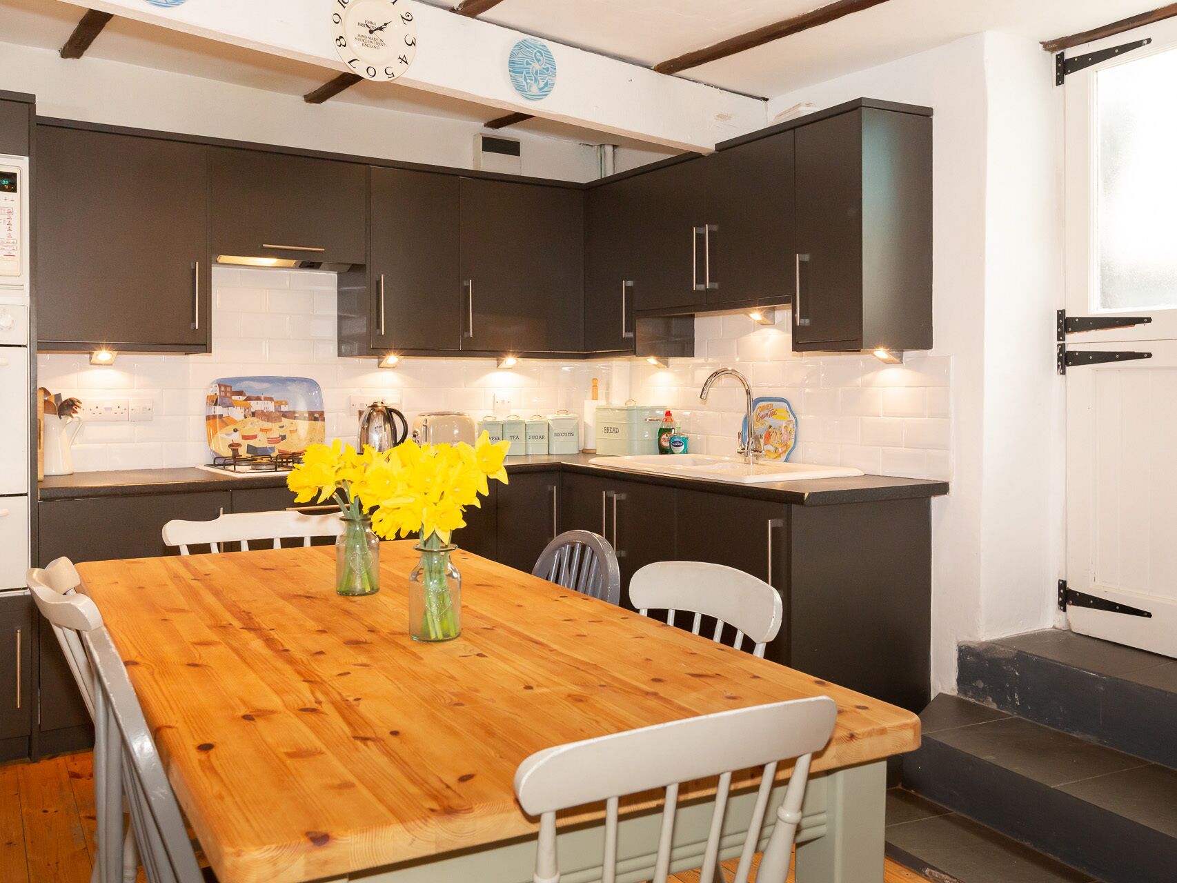 A kitchen with dark cabinets, a wooden table, and a white chair. There are vases with flowers on the table.