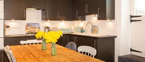 A kitchen with dark cabinets, a wooden table, and a white chair. There are vases with flowers on the table.