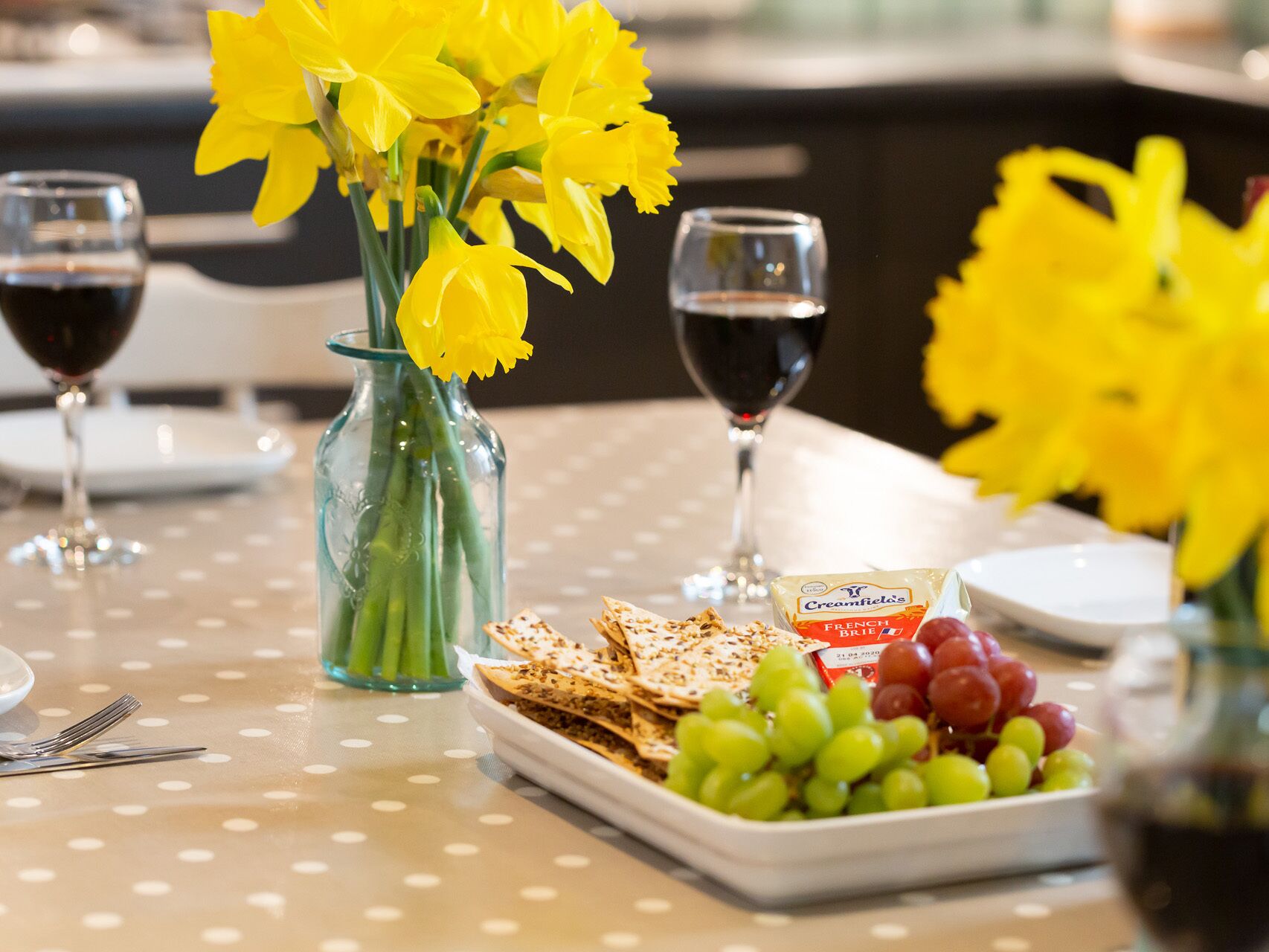 Ein Tisch mit einem Tischläufer mit Punkten, eine Vase mit gelben Blumen, eine Käse- und Crackerplatte, eine Schale mit Trauben und zwei Weingläser.