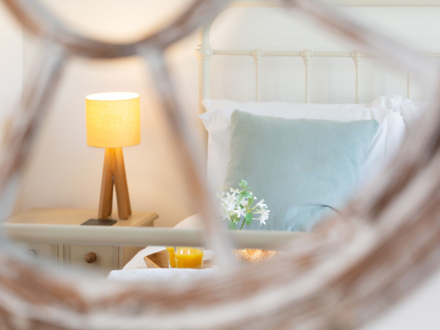 A neatly made bed with a white and light blue pillow, a bedside table with a lamp, and a small vase with white flowers.