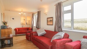 A spacious living room with a red sofa set, wooden flooring, and large windows offering a view of the outdoors.