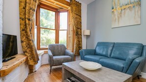 A living room with a blue sofa, a grey armchair, a wooden TV stand, and a window with curtains. - Baggergate House (York)