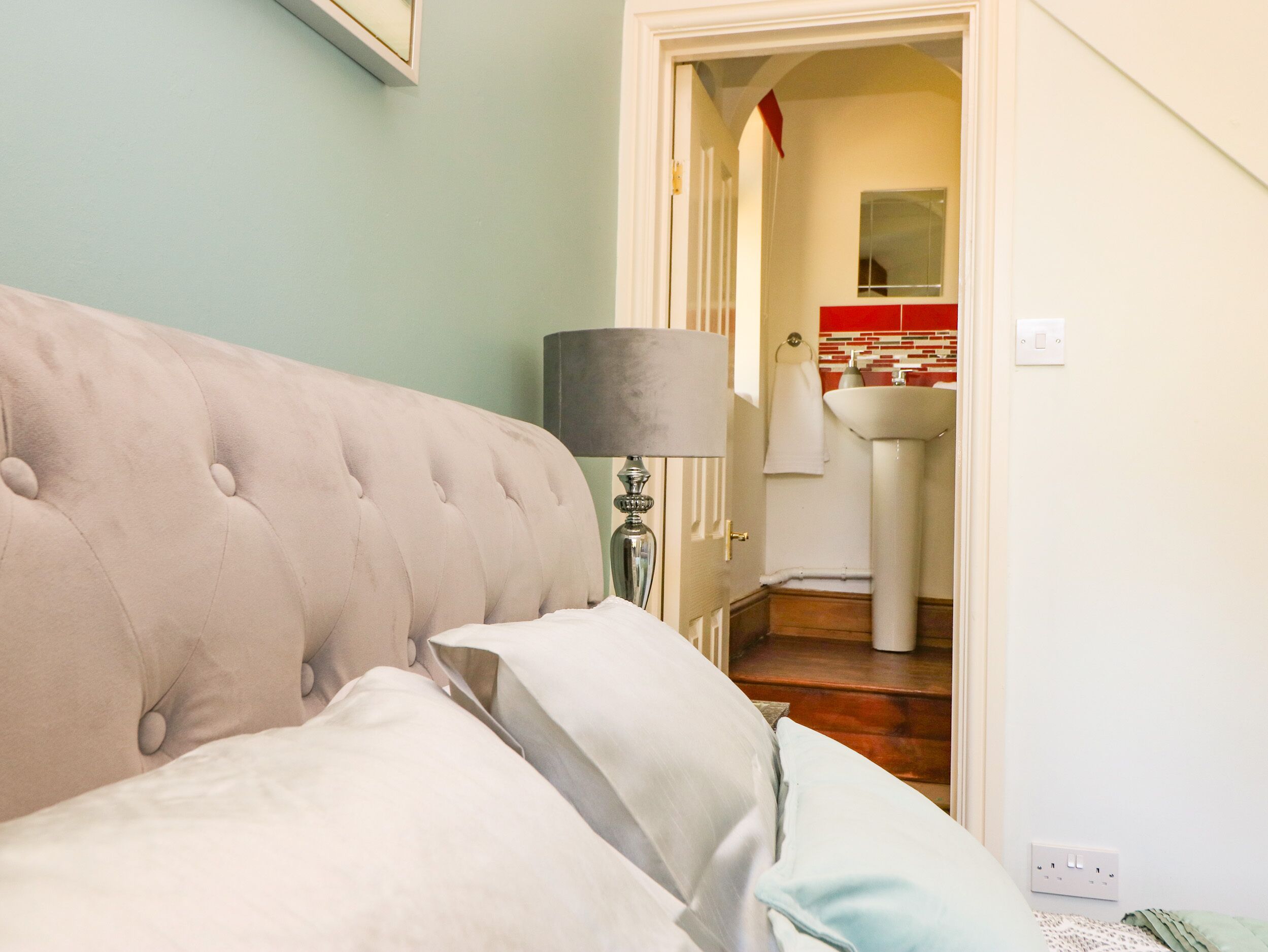 A bedroom with a tufted headboard, a bedside lamp, and a view of a bathroom with a sink and mirror.