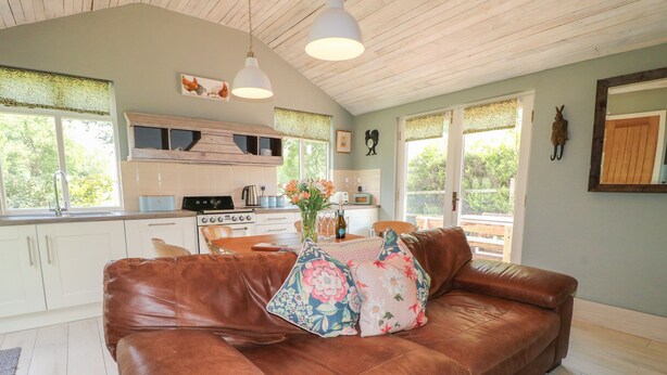 A cozy living room with a brown leather sofa, a wooden dining table, and a kitchen in the background. - The Hen House (Rugeley)