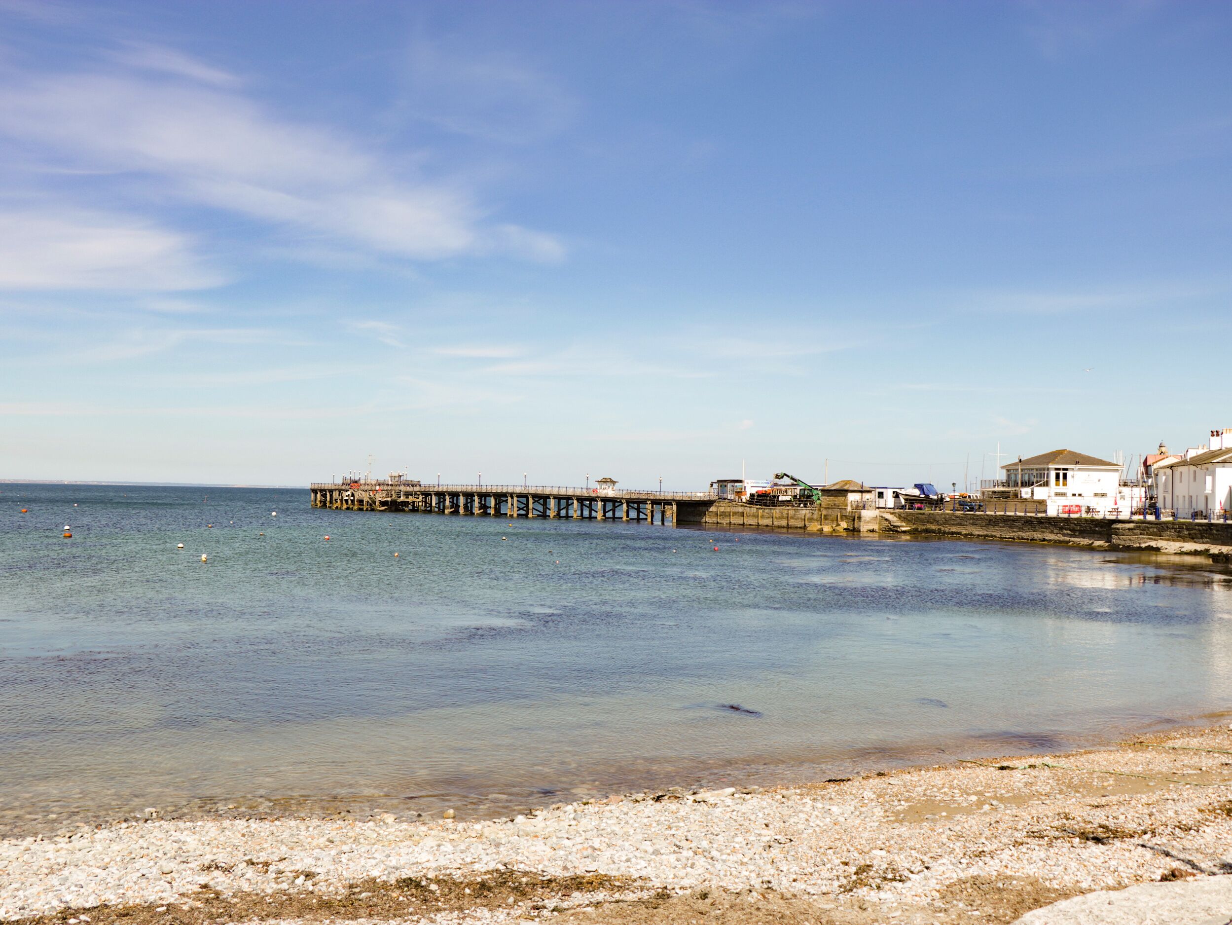 A coastal scene with a pier extending into calm waters, a sandy beach, and buildings along the shore.