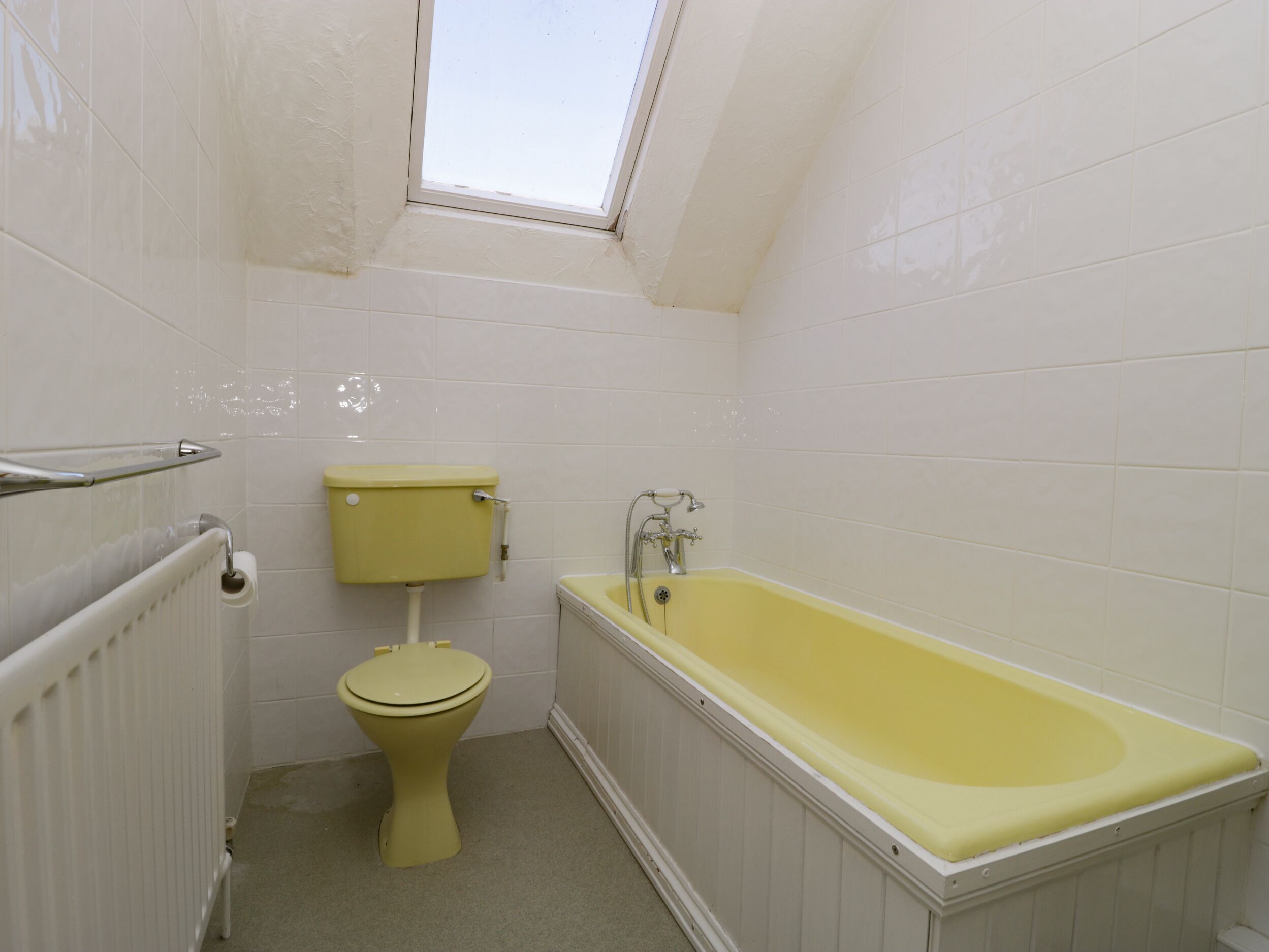 A small, narrow bathroom with white tiles, a yellow toilet, a yellow bathtub, and a skylight above the toilet.