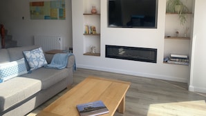 A modern living room with a sofa, coffee table, fireplace, TV, and shelves with books and decorative items.