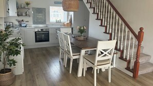 A modern kitchen with white cabinets, a dining table with chairs, a hanging light fixture, and a wooden staircase.