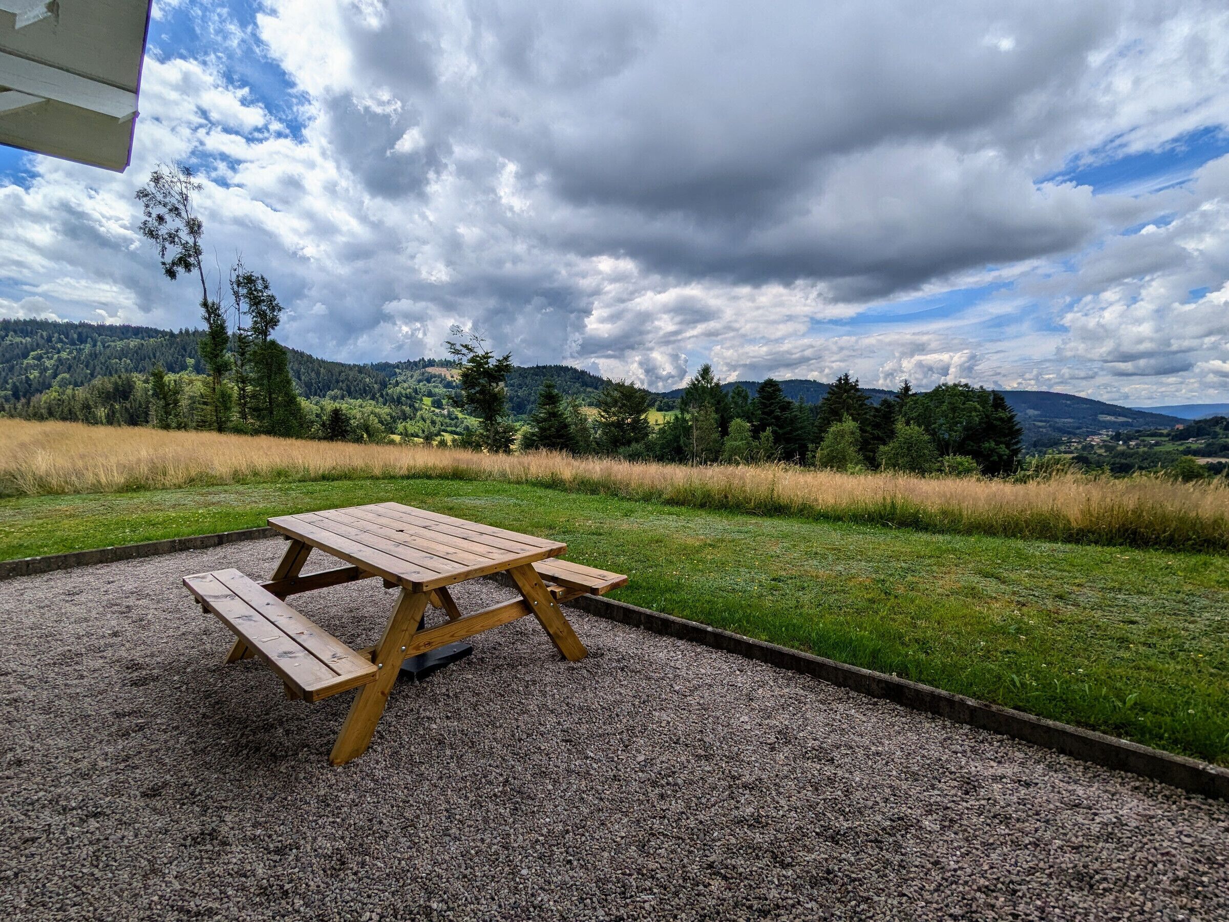 A picnic table with benches in a gravel area, surrounded by grass and trees, with a view of distant hills and a cloudy sky.