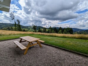 A picnic table with benches in a gravel area, surrounded by grass and trees, with a view of distant hills and a cloudy sky.