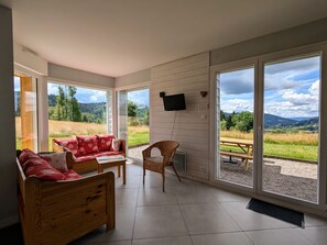 Een kamer met een houten bank, een stoel en een kleine tafel. Er is een groot raam met uitzicht op een veld en bergen.