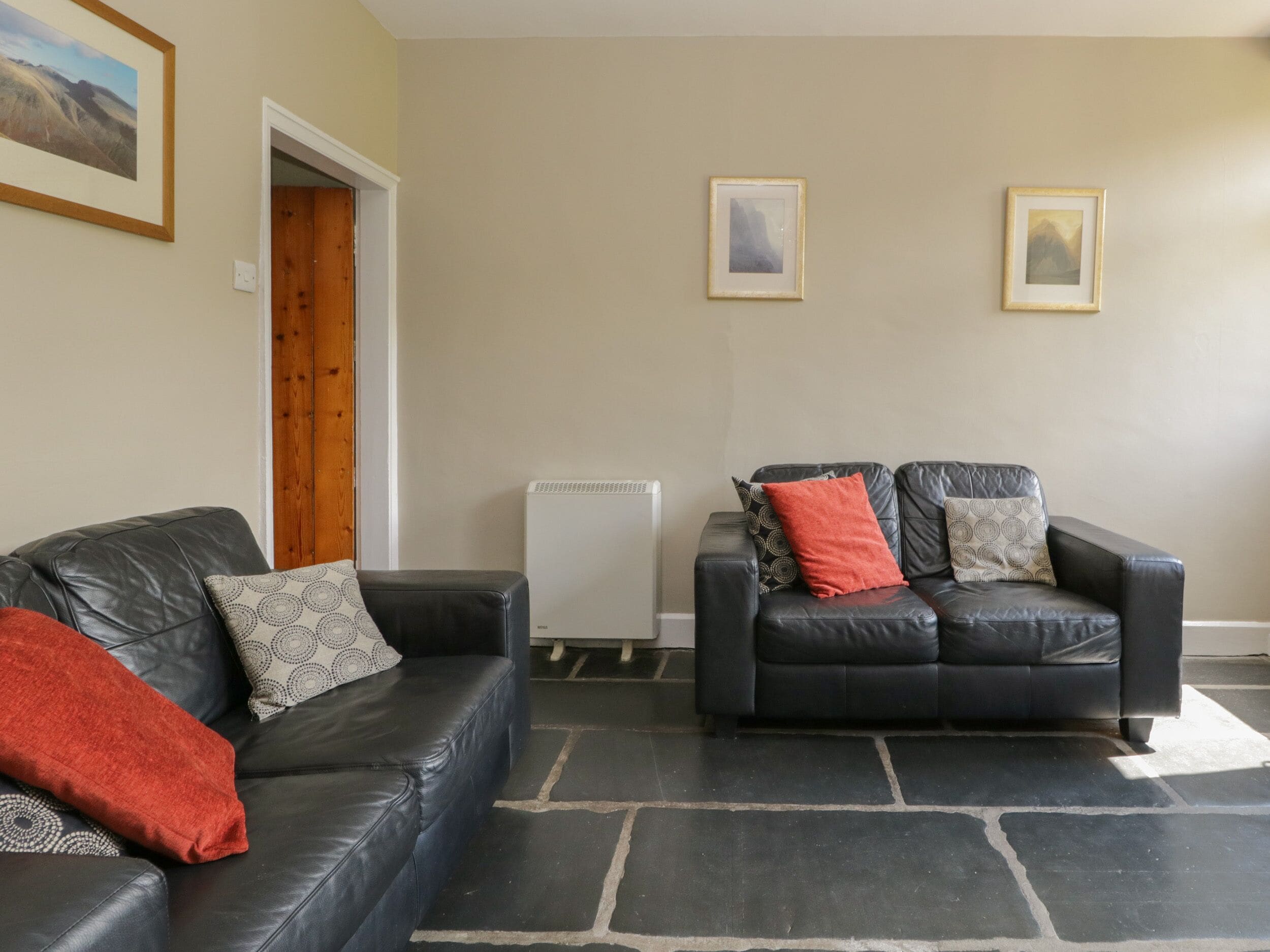 A living room with two black leather sofas, one with a red throw blanket and patterned pillows, and the other with patterned pillows. There are framed pictures on the wall, a radiator, and a wooden door.