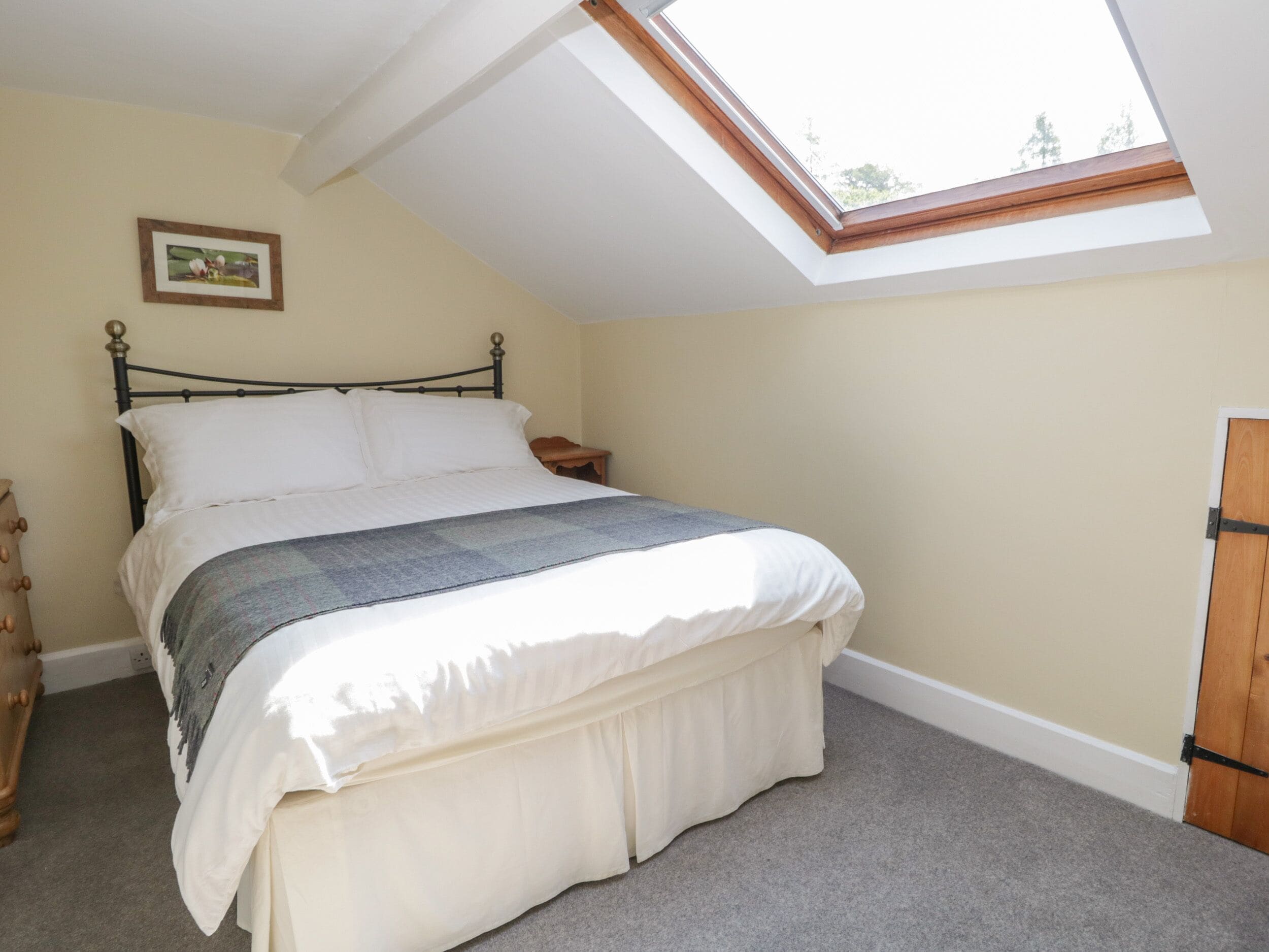 A bedroom with a skylight, a bed with a white and grey blanket, a wooden nightstand, and a dresser.