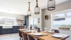 A modern kitchen with dark cabinets, a wooden dining table set for six, and a view of the sea through large windows. - Beach Haven (Dorchester)