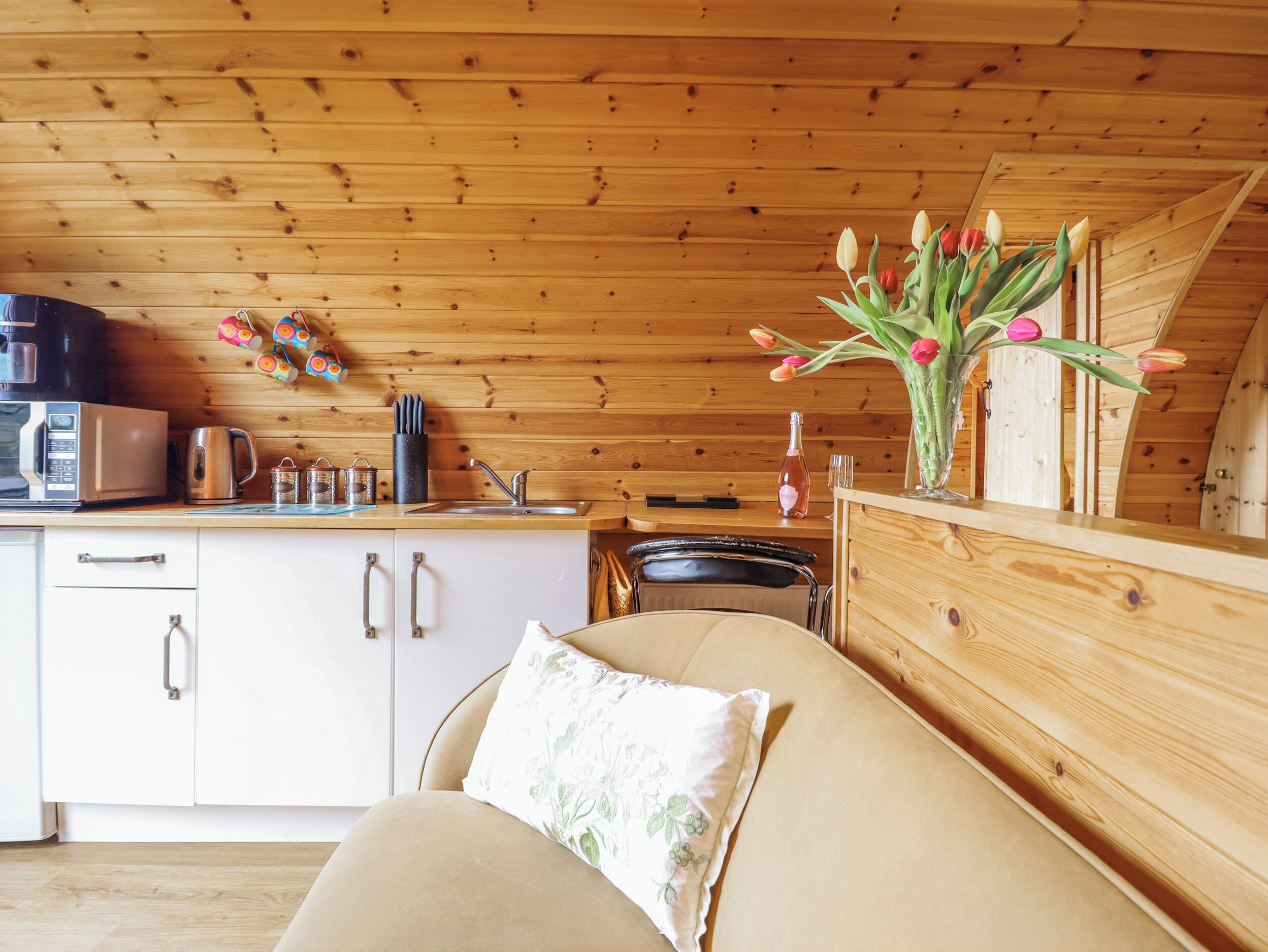Un intérieur de chalet chaleureux avec une petite cuisine, un plafond en bois et un canapé agrémenté d’un coussin à motifs floraux.