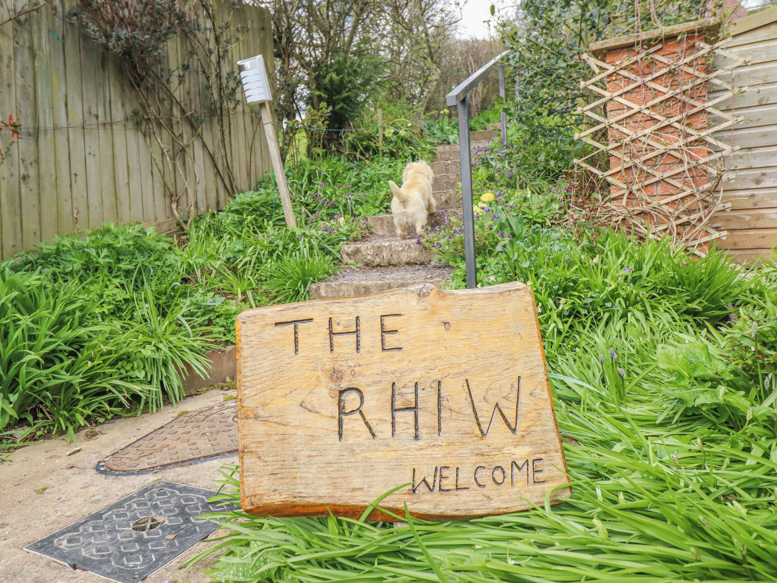 Un letrero de madera con la inscripción 'THE RHTW WELCOME' grabado, ubicado en un sendero de concreto rodeado de follaje.