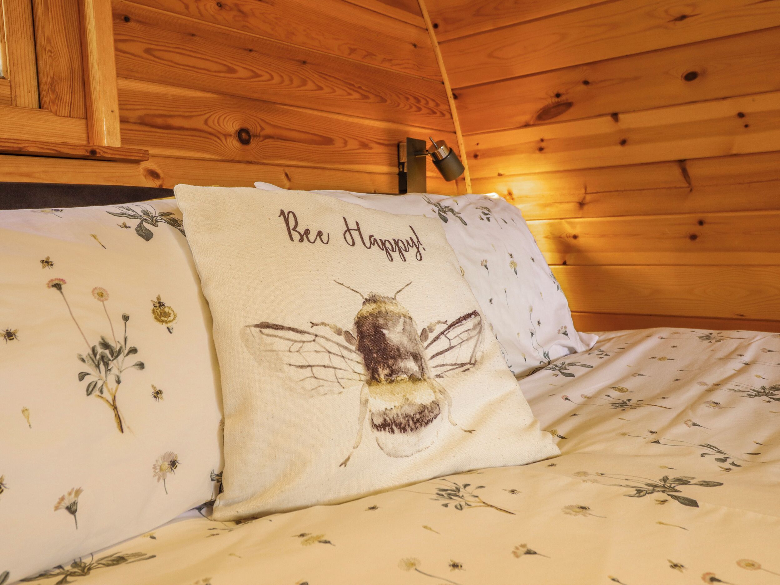 Interior de uma cabana de madeira com uma cama, roupa de cama florida e um travesseiro com ilustração de abelha e a frase 'Bee Happy!'.