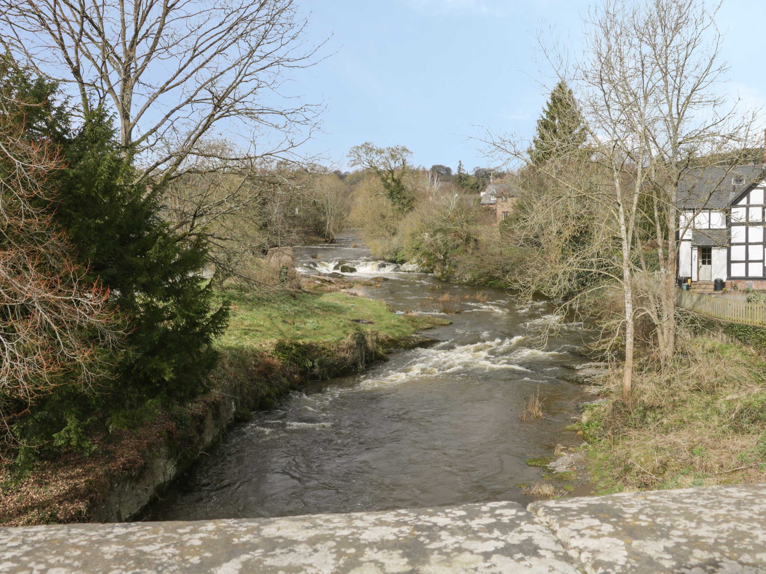 A river flowing through a rural area with a stone bridge and a traditional building on the right.