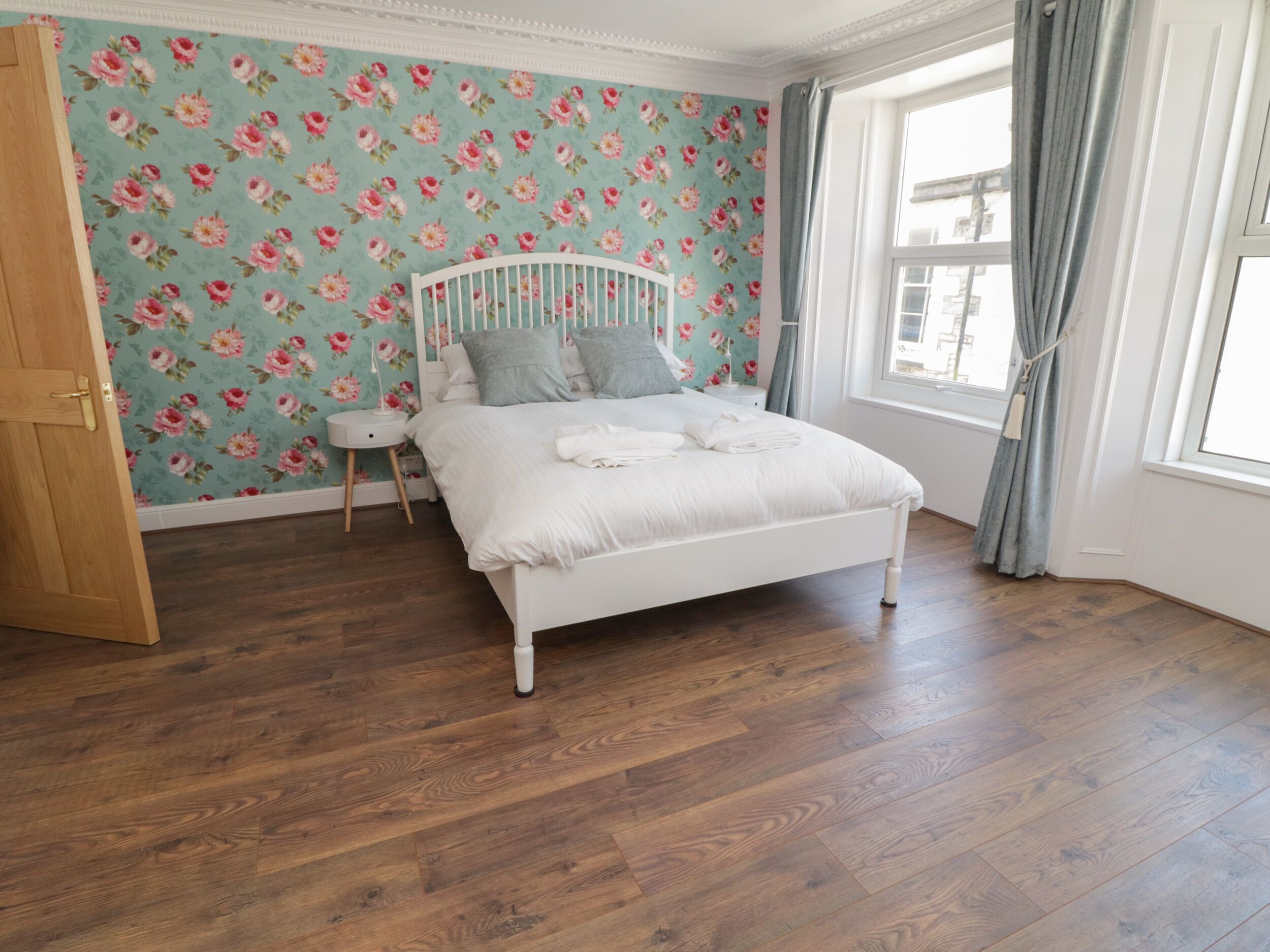 A bedroom with a floral wallpaper, a white bed, and a wooden floor.